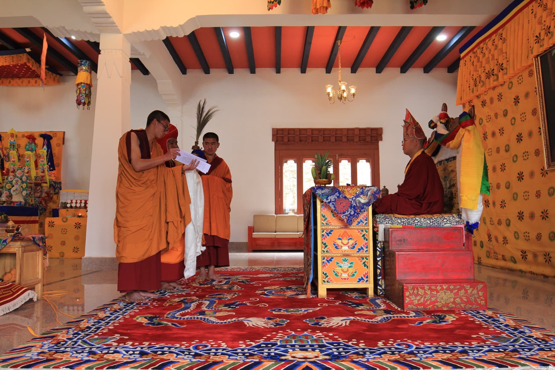 Khedrupchen Rinpoche performing an offering ritual before an ornate brocade-draped throne in a brightly painted temple hall, attended by a young monk.